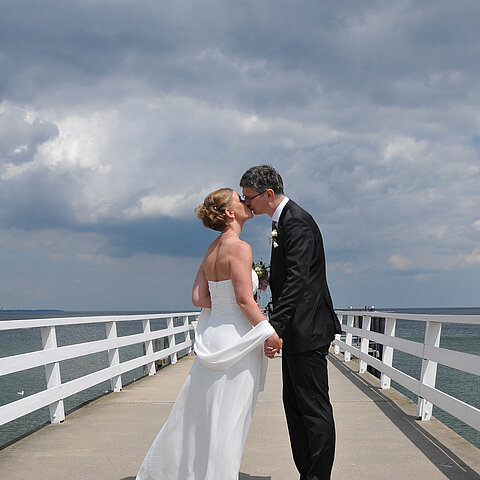 Hochzeitspaar auf der Seebrücke vor dem Maritim Hotel Timmendorfer Strand, das sich bei bewölktem Himmel einen Kuss gibt.