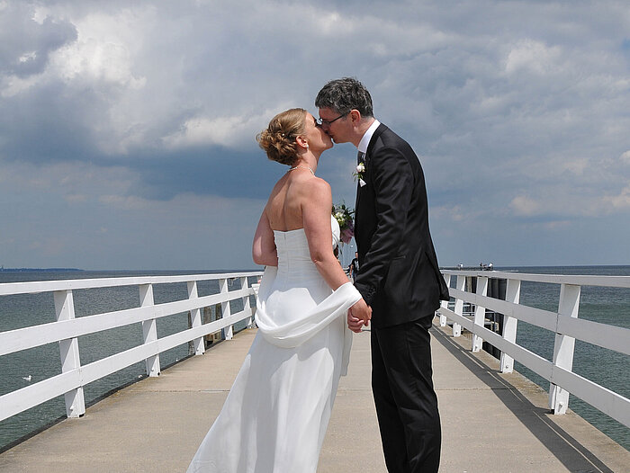 Hochzeitspaar auf der Seebrücke vor dem Maritim Hotel Timmendorfer Strand, das sich bei bewölktem Himmel einen Kuss gibt.