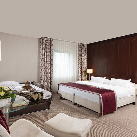 Comfort Familienzimmer im Maritim Hotel Bonn mit großem Doppelbett, ausziehbarem Sofa, Sessel und Blumenarrangement.