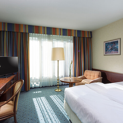 Helles Comfort Zimmer im Maritim Hotel Köln mit Einzelbett, Schreibtisch, Fernseher, Sessel und großen Fenstern mit Vorhängen