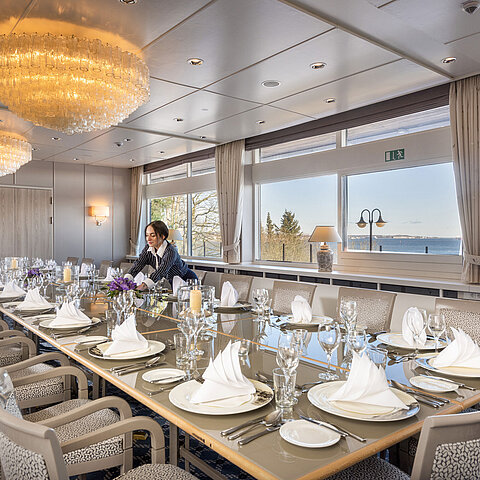 Salon Oslo im Maritim Hotel Kiel, langer festlich gedeckter Tisch mit Meerblick, Kronleuchter, Personal richtet den Raum.