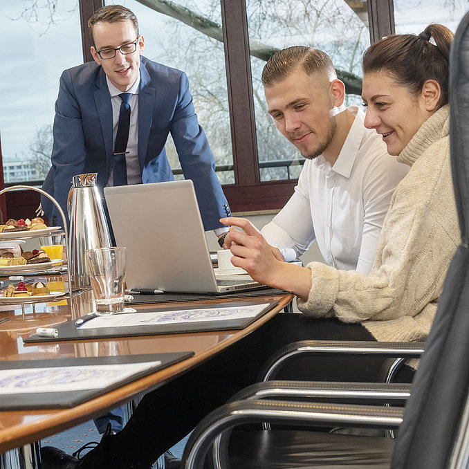 Team in einer Besprechung im Boardroom Maritim Köln, mit Kaffee und Snacks auf dem Tisch, während sie am Laptop arbeiten.