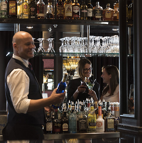 Fröhliche Gäste genießen Cocktails an der Bar des Maritim Hotel Dresden, serviert von einem freundlichen Barkeeper.