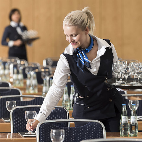 Eine Servicekraft bereitet einen Konferenzraum vor, trägt ein Tablett mit Gläsern und stellt Wasserflaschen bereit.