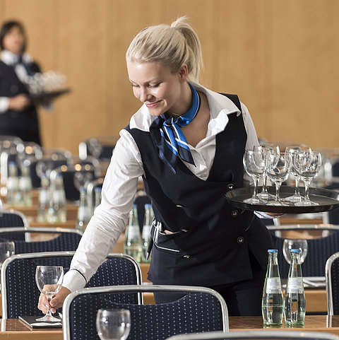 Eine Servicekraft bereitet einen Konferenzraum vor, trägt ein Tablett mit Gläsern und stellt Wasserflaschen bereit.
