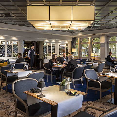 Elegantes Restaurant mit Gästen, die speisen und entspannen. Moderne Einrichtung und große Fenster mit Ausblick.