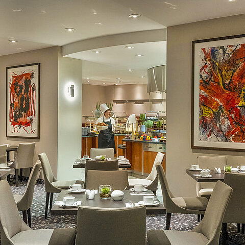 Restaurant im Maritim proArte Hotel Berlin mit moderner Kunst an den Wänden, gedeckten Tischen und einem Koch am Buffet.