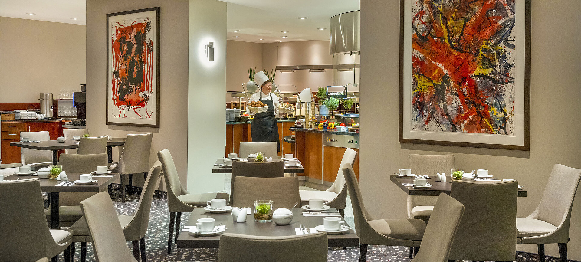 Restaurant im Maritim proArte Hotel Berlin mit moderner Kunst an den Wänden, gedeckten Tischen und einem Koch am Buffet.