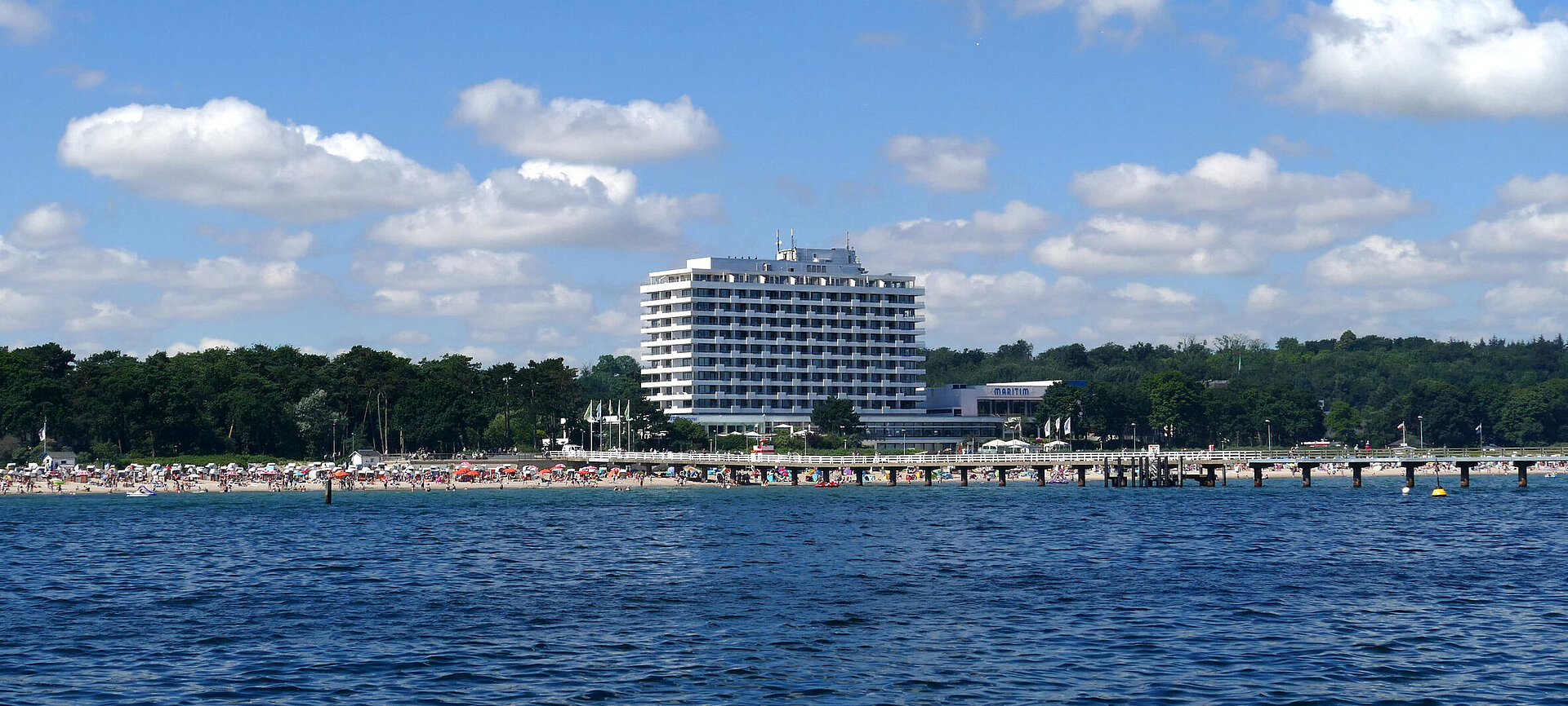 Maritim Seehotel Timmendorfer Strand mit Seebrücke, Strandbesuchern und blauem Himmel im Sommer.