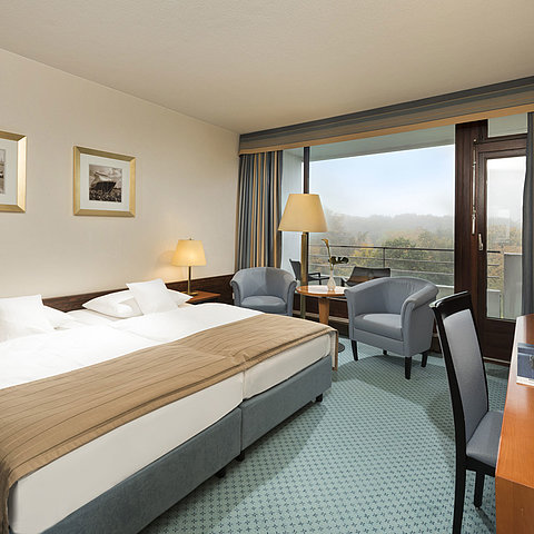 Classic Zimmer im Maritim Hotel Kiel mit großem Doppelbett, Sitzbereich, Schreibtisch und Balkon mit Blick ins Grüne.