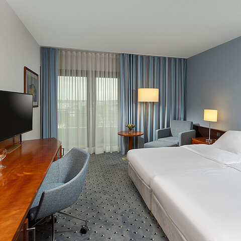 Classic Doppelzimmer im Maritim Darmstadt mit Schreibtisch, Flachbildfernseher, Sitzecke und großen Fenstern mit Vorhängen.