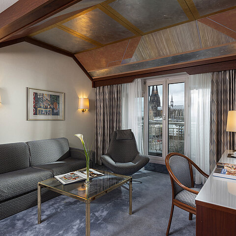 Junior Suite im Maritim Hotel Köln mit Blick auf den Kölner Dom, stilvollem Sofa, Sessel und Schreibtisch mit Lesematerial.